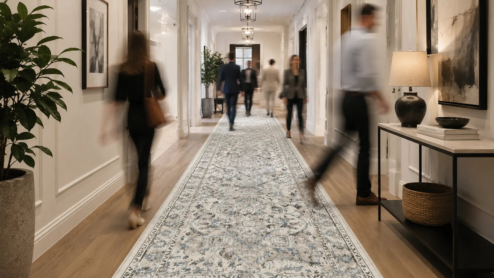 A long patterned area rug in a light blue, white, and beige traditional design is shown in a real hallway with people walking through it. The hallway has white walls, wood floors, framed wall art.