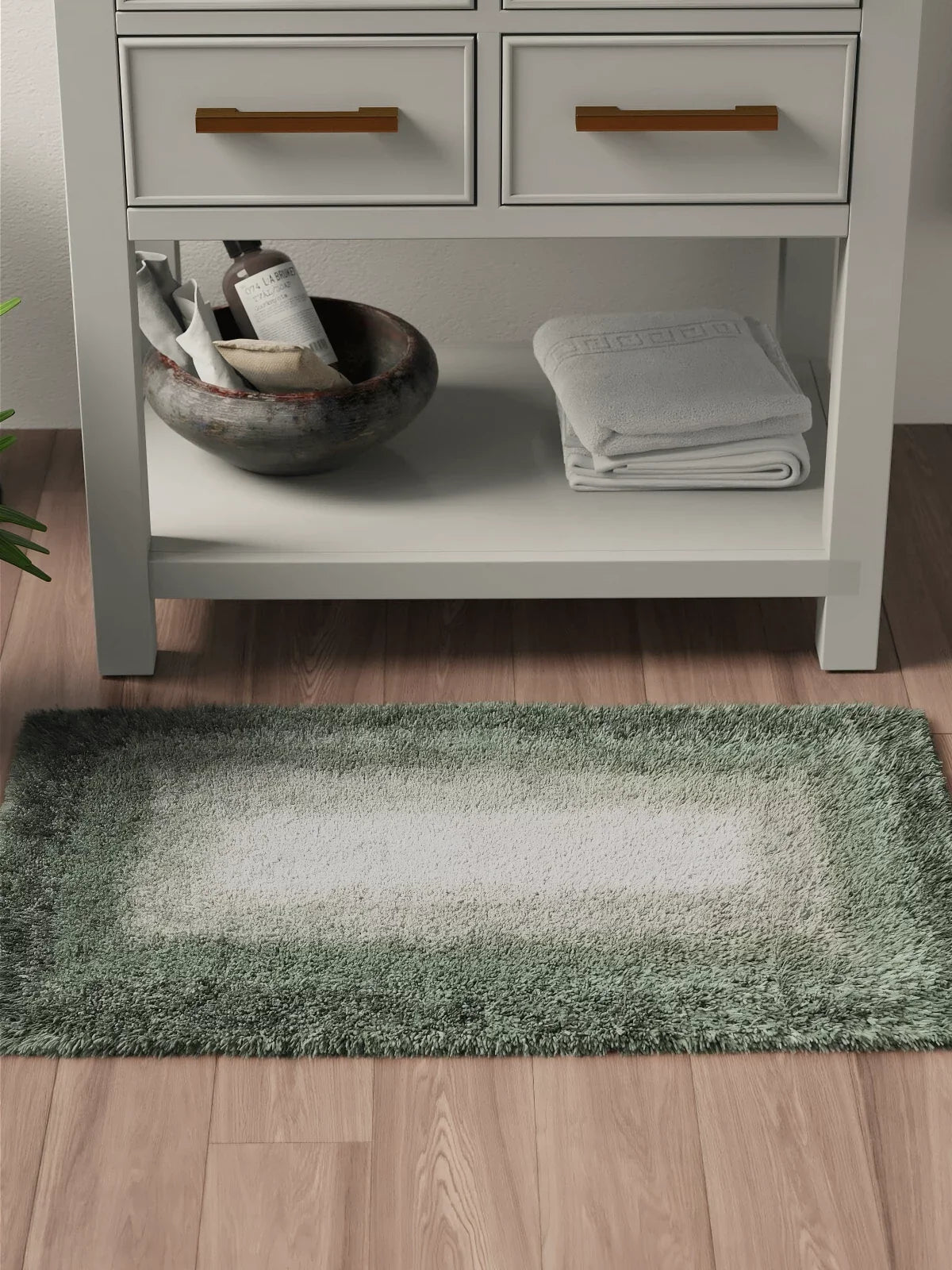 Soft 2x3 area rug in gradient green tones placed in front of a white bathroom vanity with neatly folded towels and spa essentials on a wooden floor.