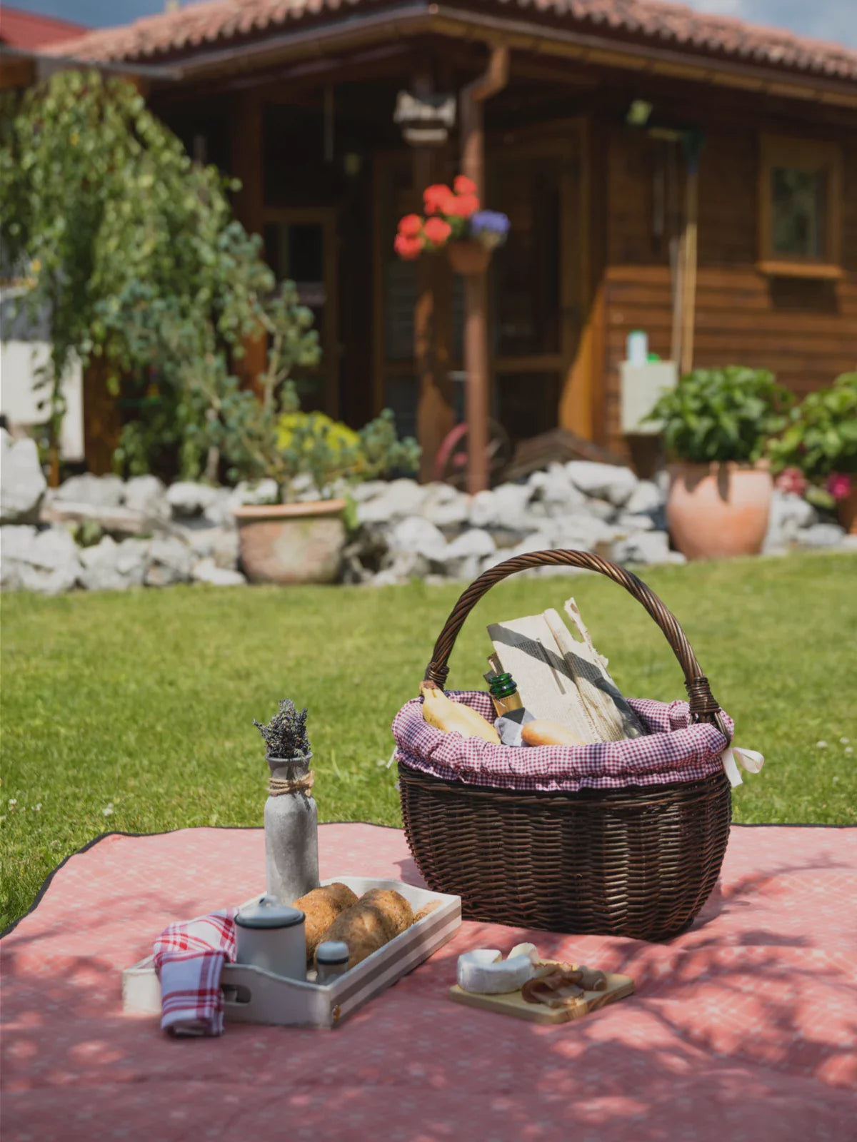 Cozy outdoor picnic setup on a pink 4x6 rug, featuring a wicker basket filled with bread and wine, a tray with fresh pastries, and a charming wooden house in the background.