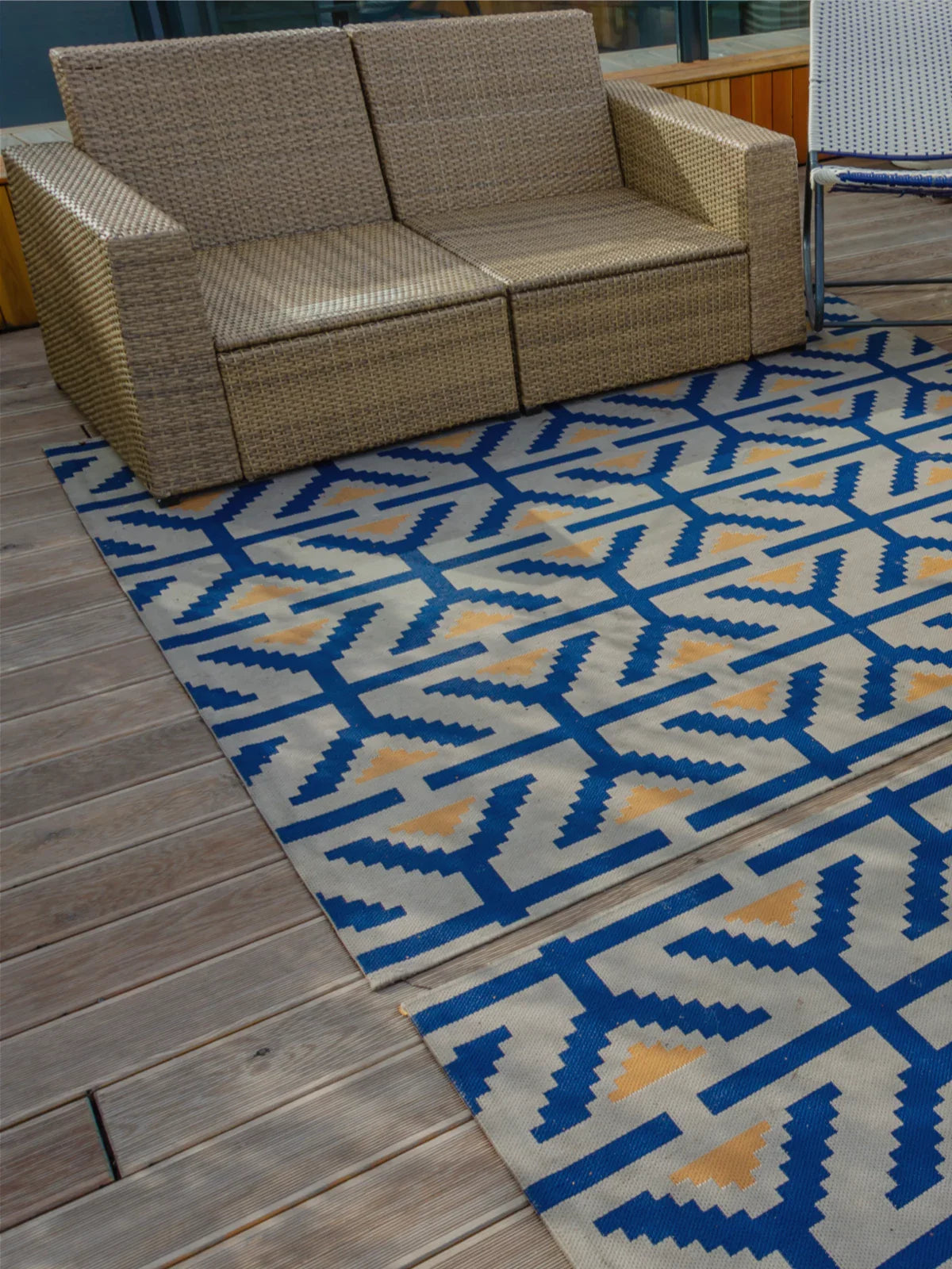 Stylish 8x10 outdoor rug with a bold blue and beige geometric pattern, placed on a wooden deck alongside a woven rattan sofa and a lounge chair, creating a cozy outdoor retreat.