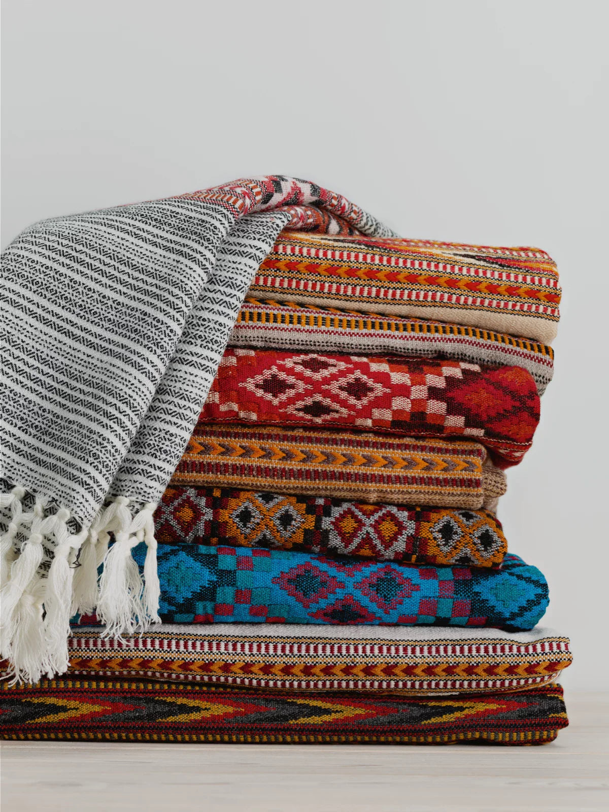 Stack of colorful handmade rugs with intricate patterns, showcasing traditional craftsmanship and rich cultural heritage. A woven black-and-white throw with tassels is draped over the top, adding contrast to the vibrant designs.