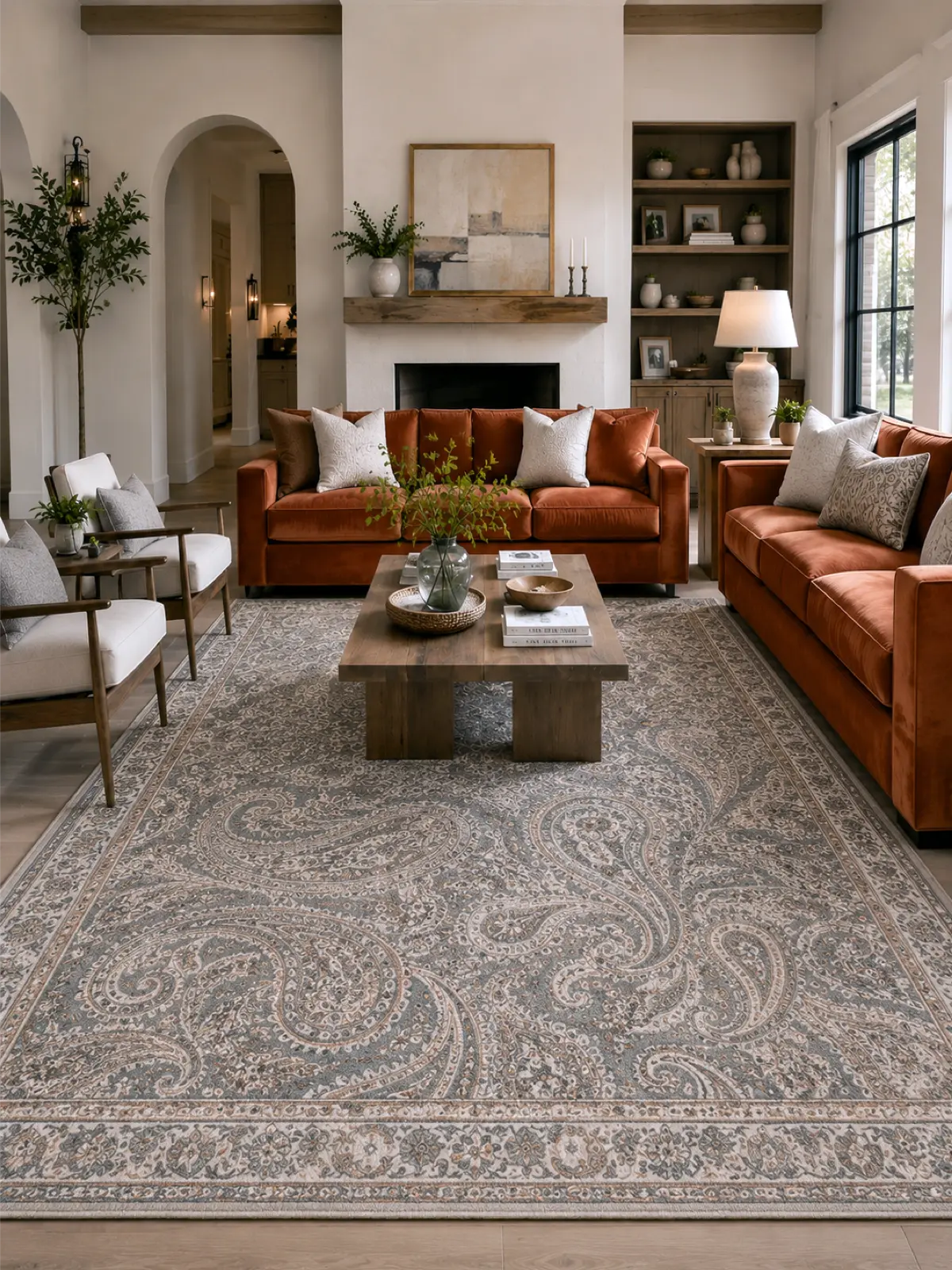 A large traditional-style area rug with a gray and beige paisley pattern, shown in a styled living room with orange-brown leather sofas, a wooden coffee table, and neutral decor.