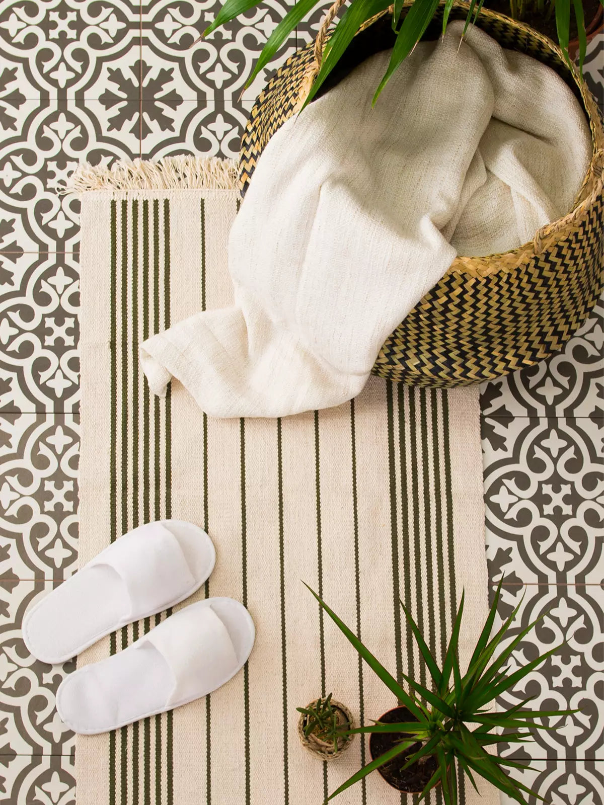 Minimalist bathroom setup featuring a striped, washable rug on patterned tiles, complemented by white slippers, a woven basket, and greenery.