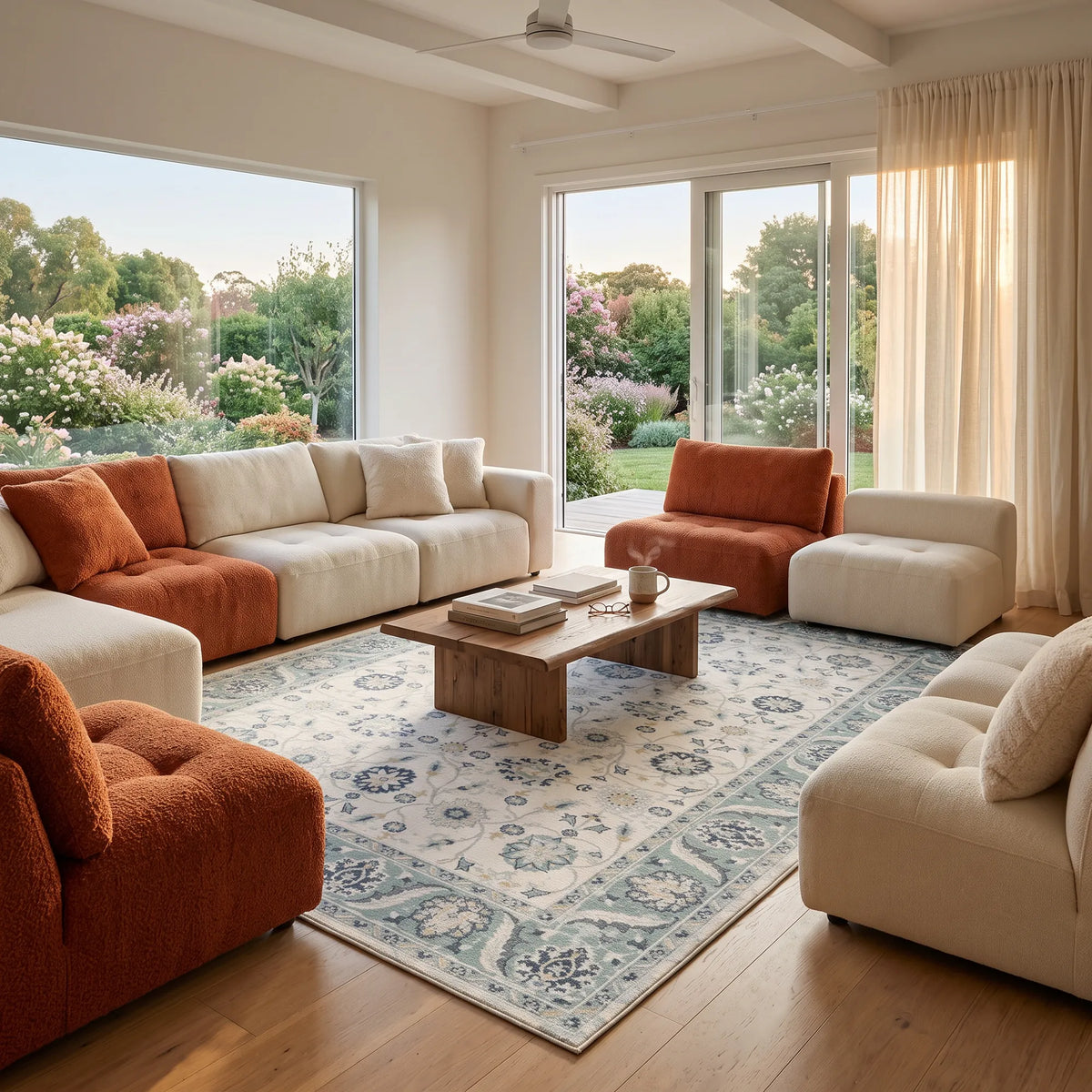 Ein modernes Wohnzimmer mit cremefarbenen und rostorangefarbenen Ecksofas, die um einen hölzernen Couchtisch auf einem blau gemusterten Teppich angeordnet sind, mit großen Fenstern und Blick auf einen Garten.