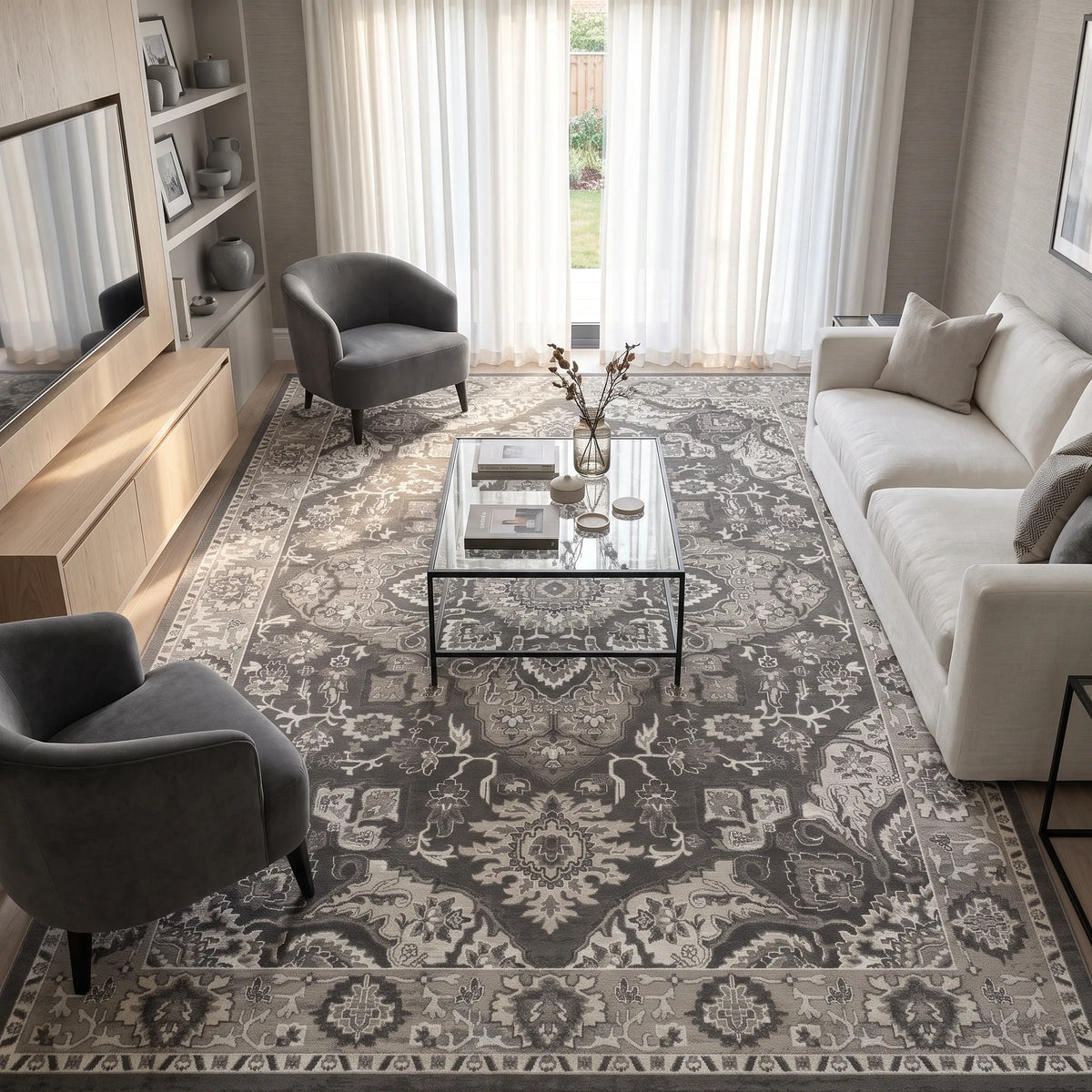 Ein großer Teppich im traditionellen Stil in Grau- und Cremeweißtönen, arrangiert in einem modernen Wohnzimmer mit einem Couchtisch mit Glasplatte, einem hellgrauen Sofa, zwei grauen Sesseln und einem Medienmöbel aus Holz.