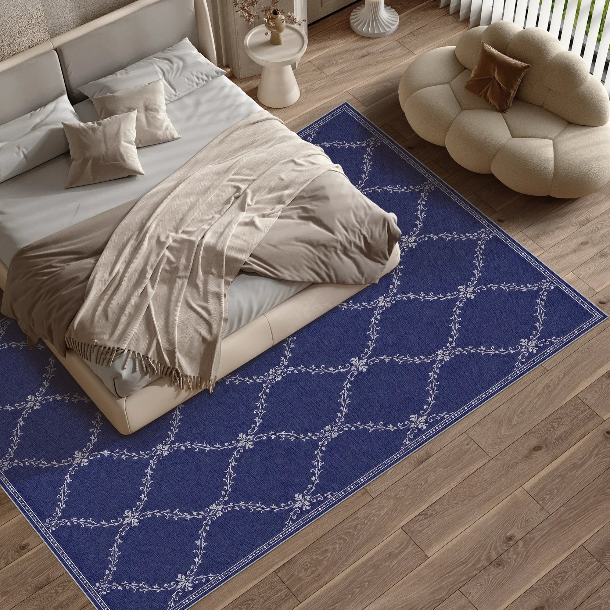 A rectangular blue lattice area rug with a white floral trellis pattern, shown in a styled bedroom setting next to a bed and a cream cloud-shaped pouf on a wood floor.